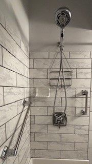 A tiled shower featuring a handheld shower head, a chrome caddy, a soap dish, and two installed grab bars for accessibility.