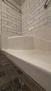 A white, low-profile shower base with a built-in seat, set against white subway-tiled walls and a dark gray tile floor.