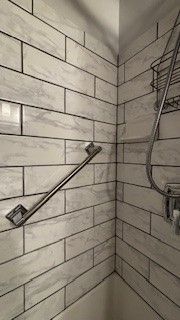 A diagonal metal grab bar installed on light-colored marble-patterned subway tile walls in a shower corner.