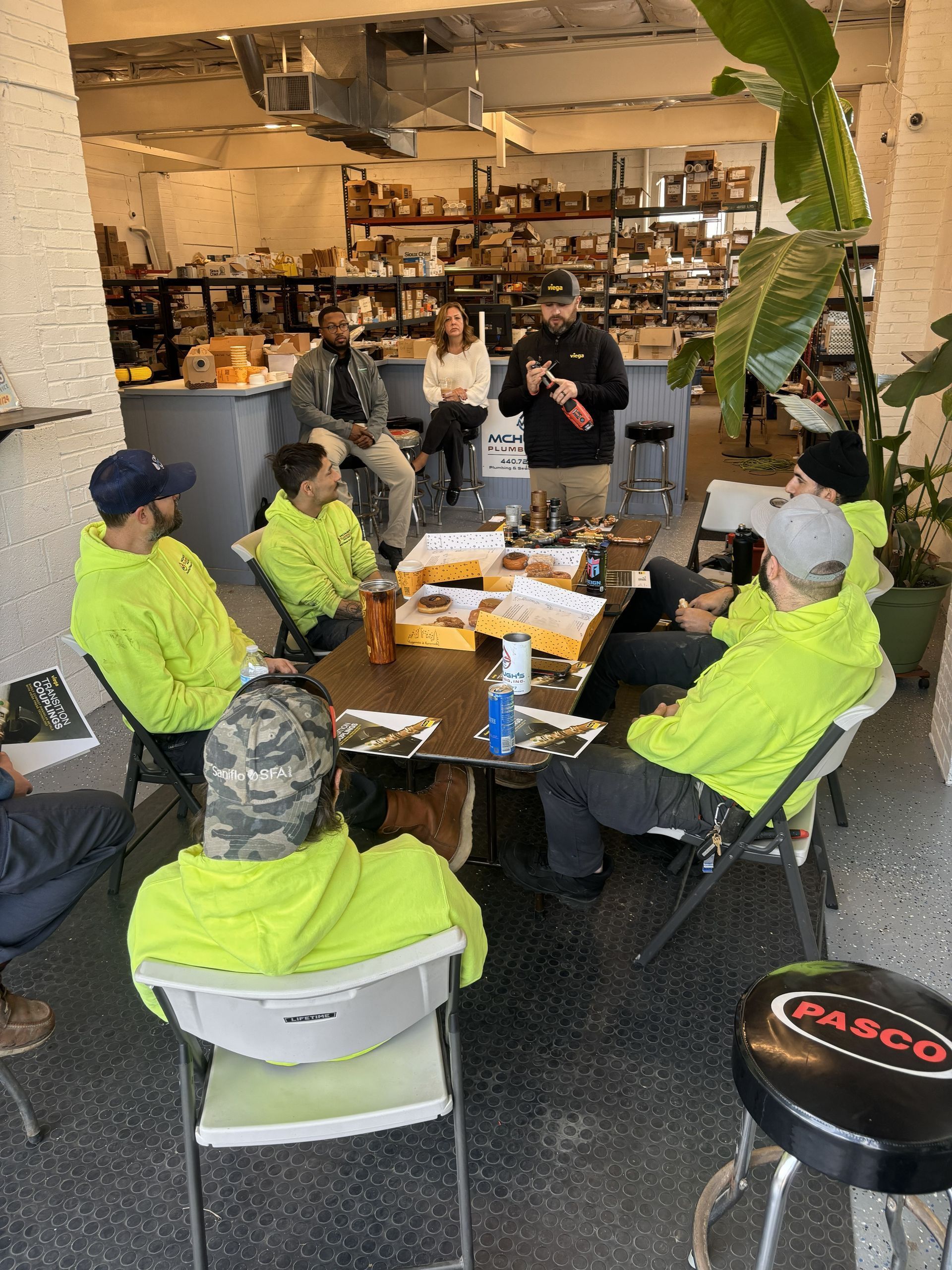 Group of people in neon green hoodies at a table, some eating. A man stands, speaking. Workshop or meeting setting.