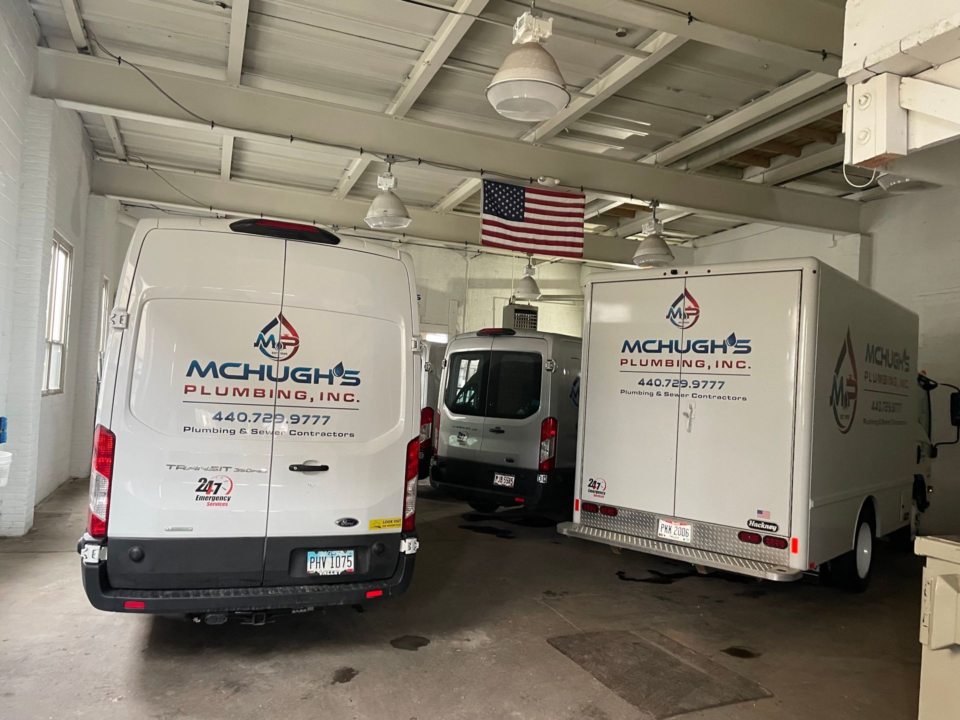 Three white plumbing service vans parked in a garage. The vans have the company logo and text on the sides. An American flag hangs inside.