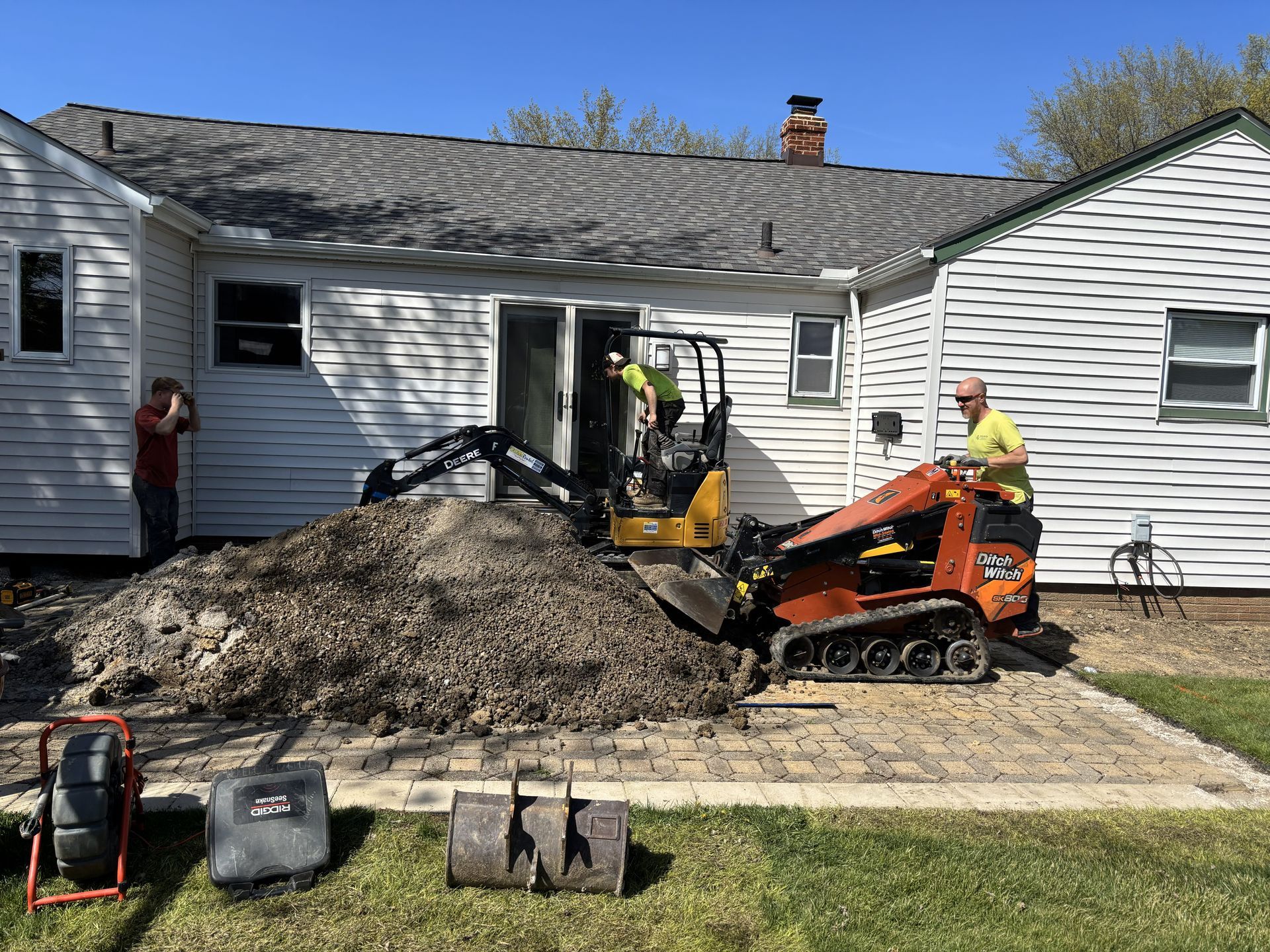 Construction site: men operating mini-excavators near a house. Dirt pile, patio, and tools are visible.