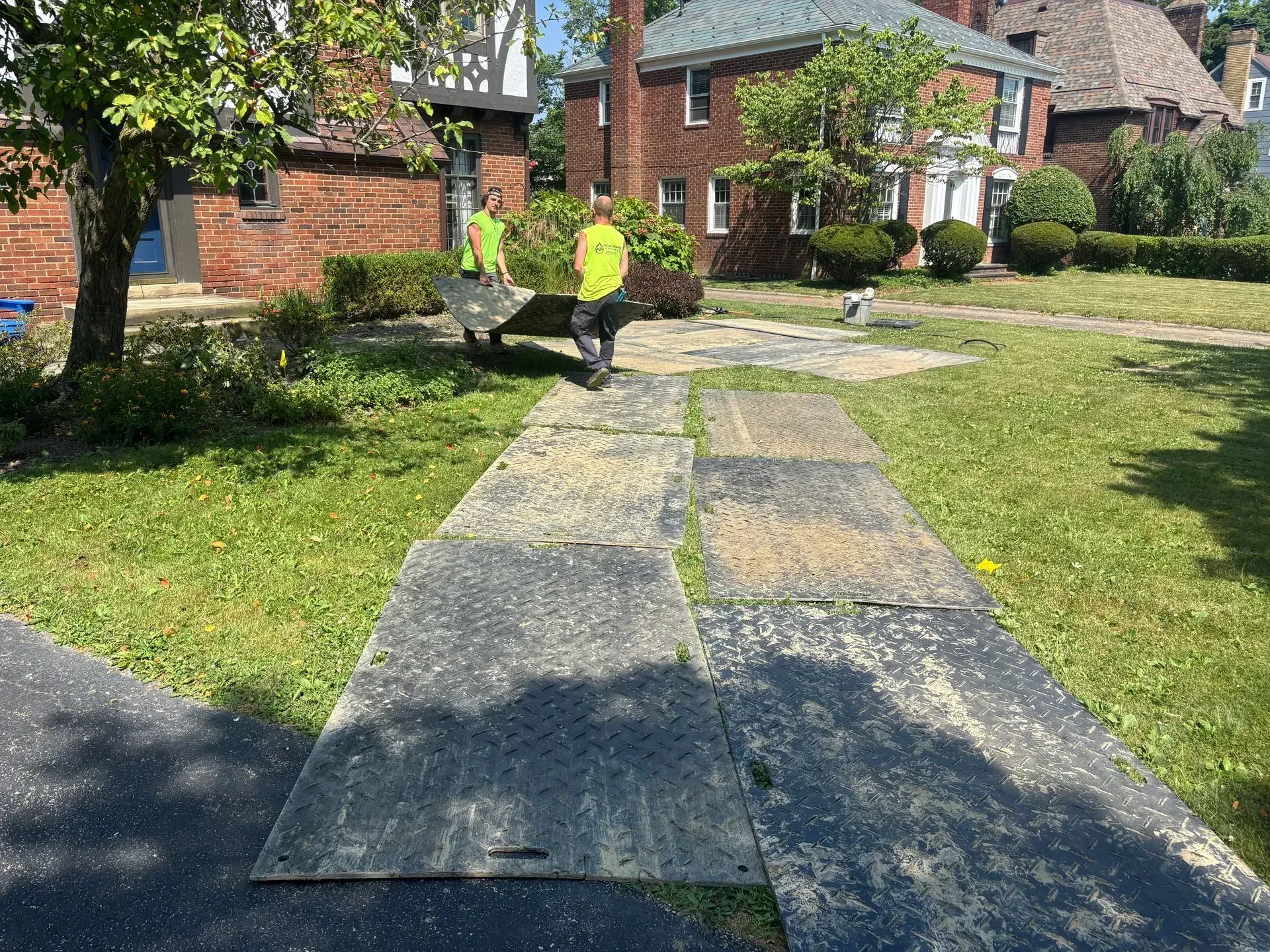 Workers carrying a dark panel over a temporary walkway of panels on grass near brick buildings.