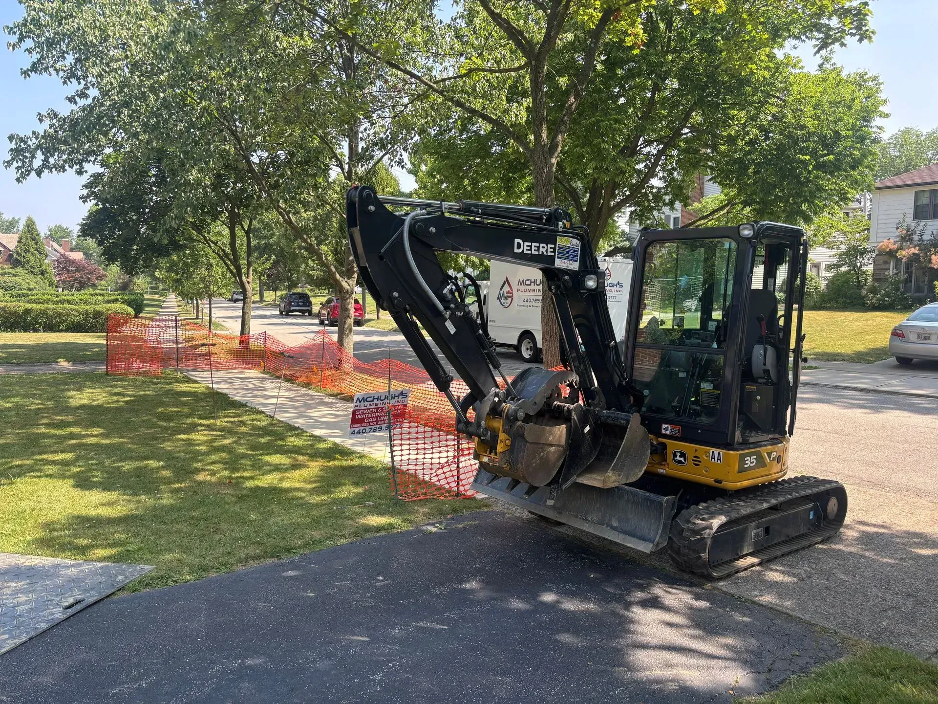 Small excavator on asphalt driveway next to orange safety fencing. Trees, suburban street visible.