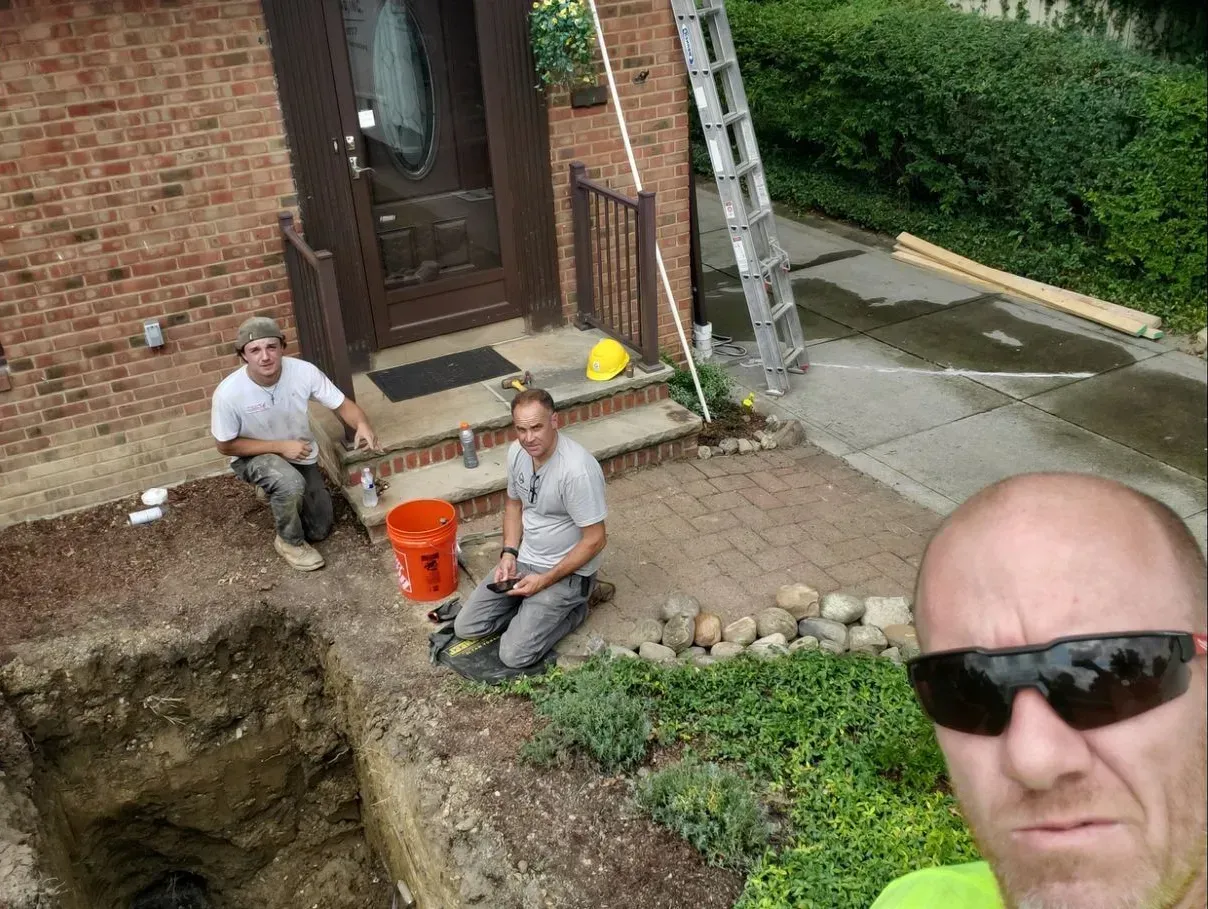 Three workers near a brick house; one takes a selfie. A hole in the ground and a ladder are visible.