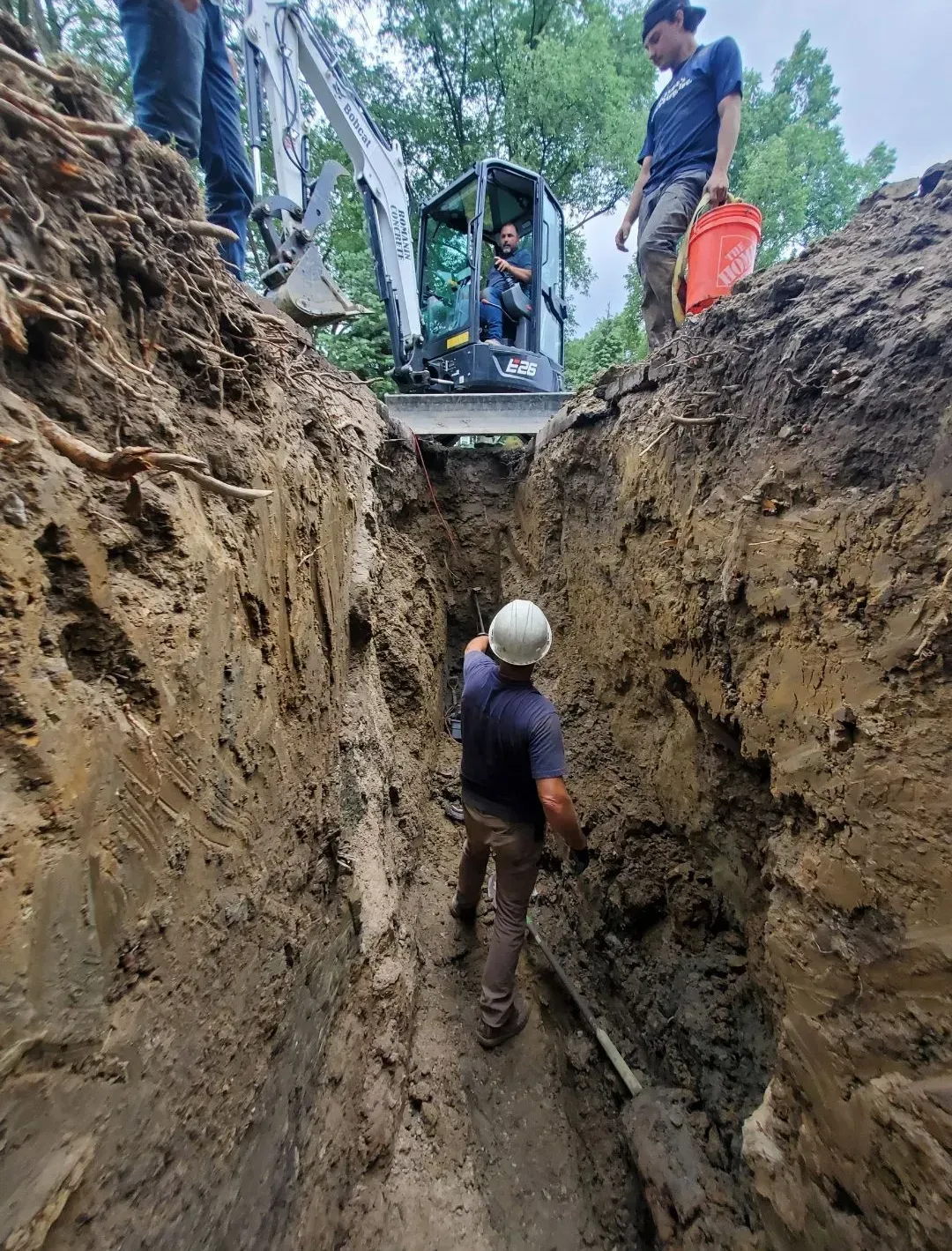 Workers excavating a trench with an excavator and hand tools outdoors; cloudy day.