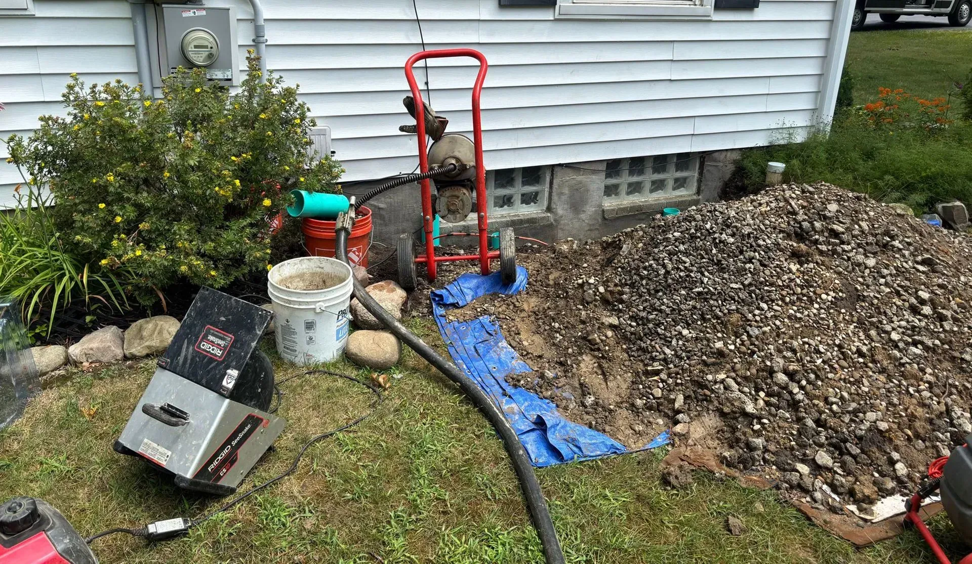 Construction site next to a white house with tools, gravel, and a blue tarp on the grass.