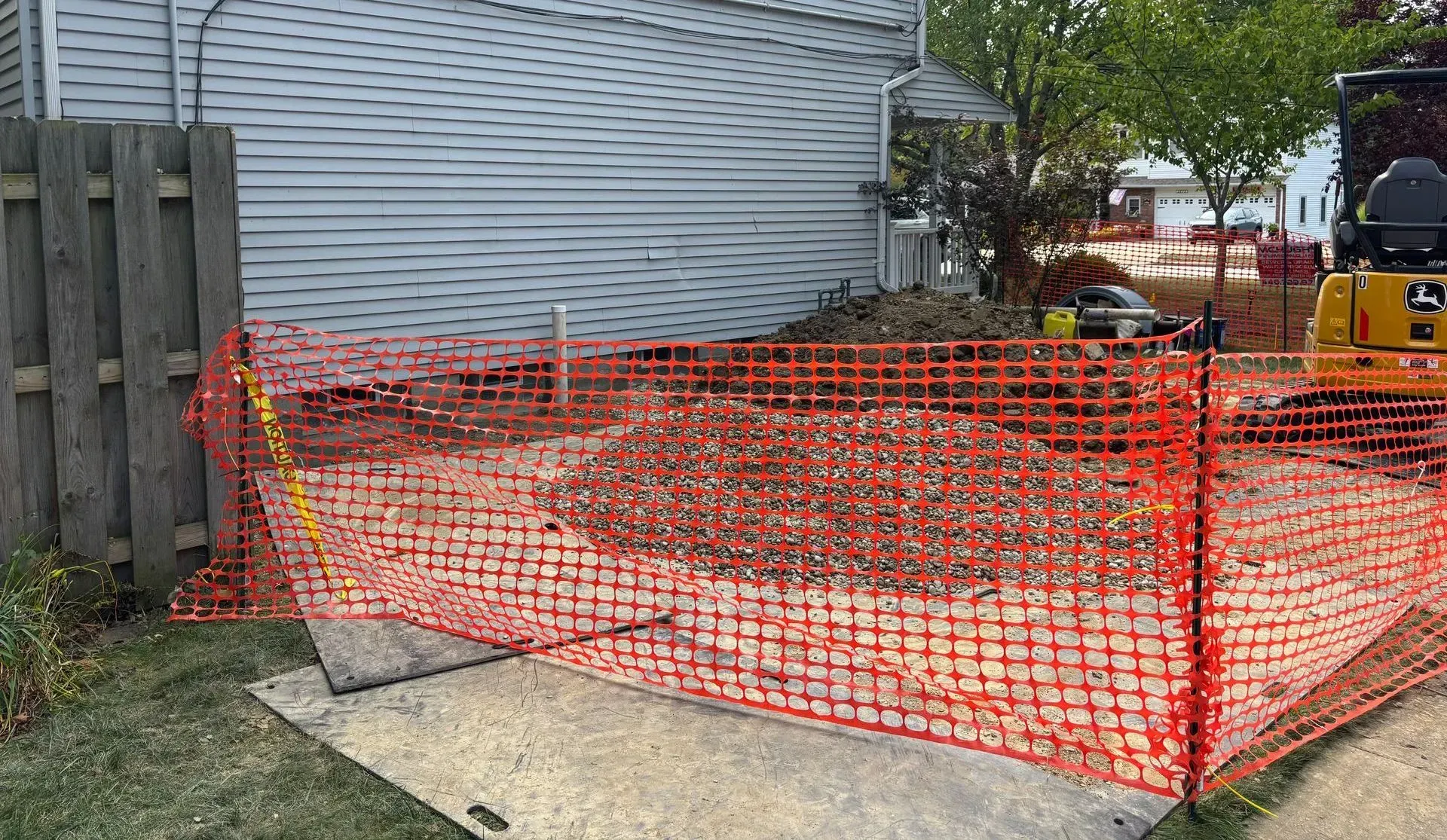 Orange safety fencing surrounds a pile of debris and small excavator. Backyard setting.