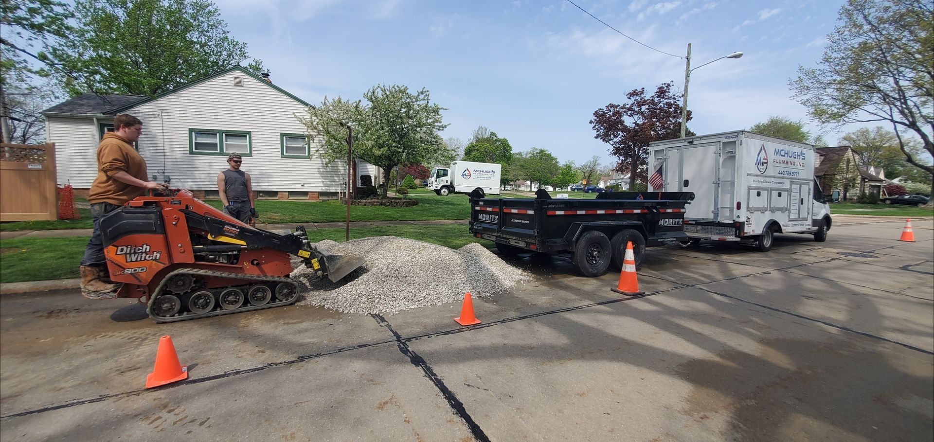 A person operates a small construction vehicle, moving gravel on a street. A trailer and truck are parked nearby.
