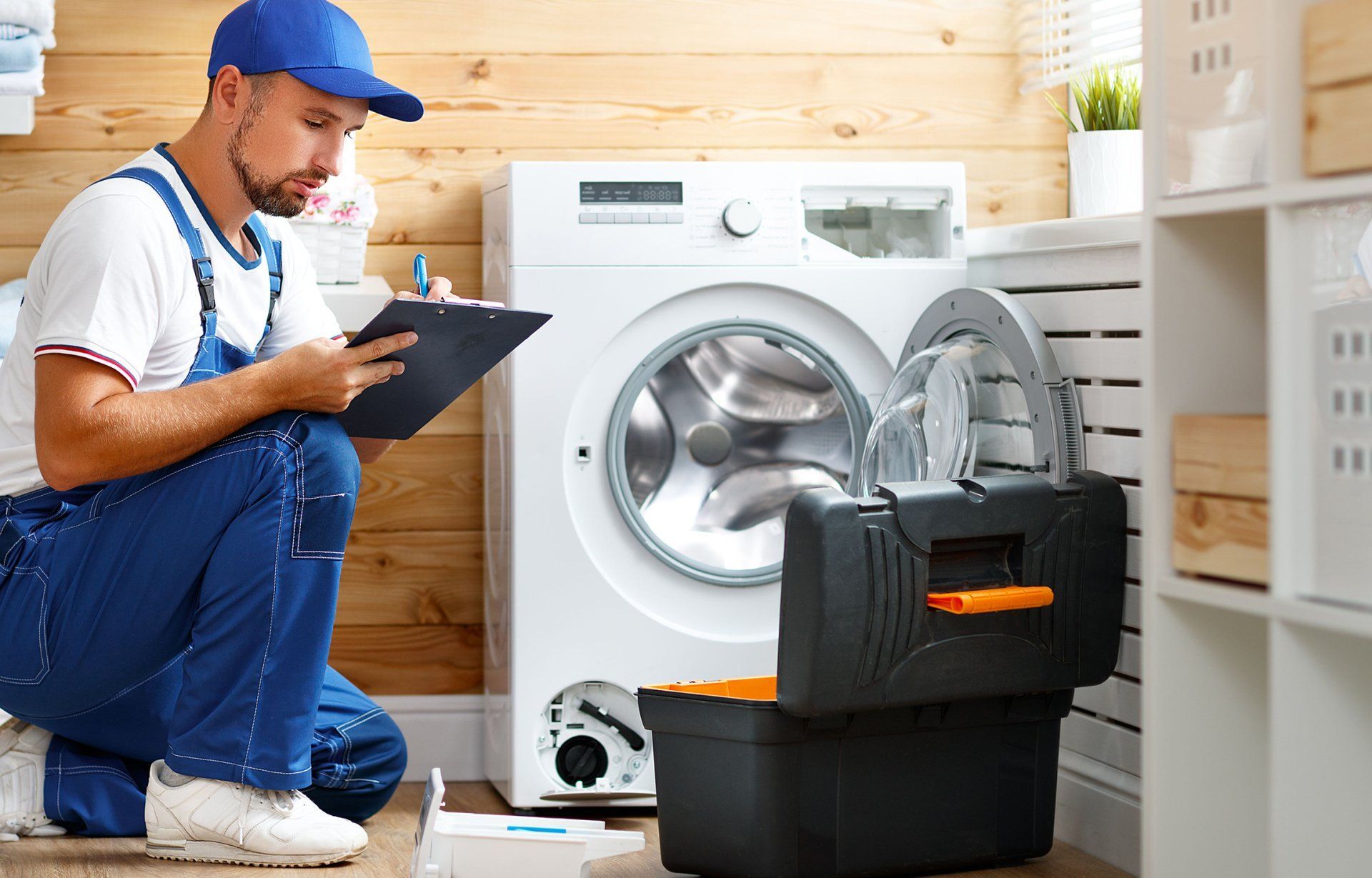 A repairman in blue overalls kneels by a washing machine, taking notes from an open toolbox in a laundry room.