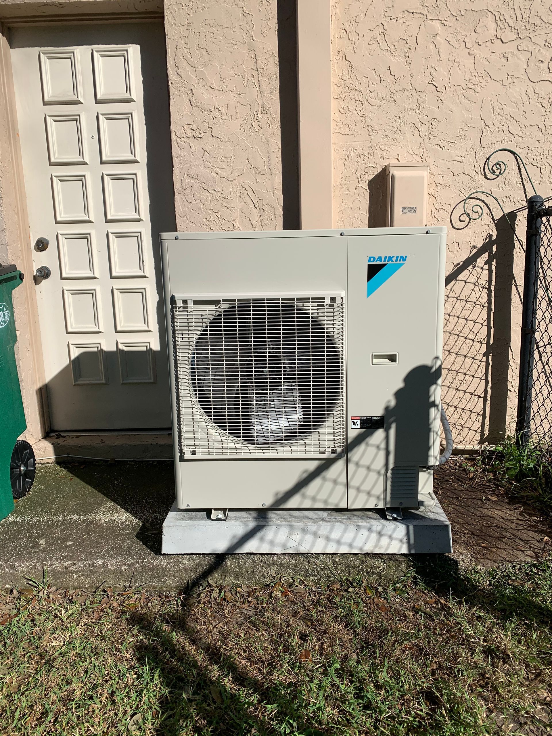 A large air conditioner is sitting outside of a building next to a trash can.