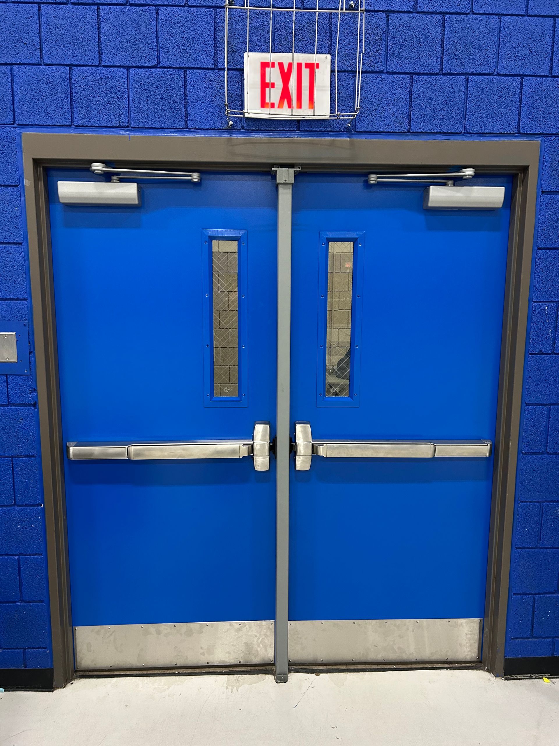 Blue double doors with silver hardware and an EXIT sign above against a blue brick wall.