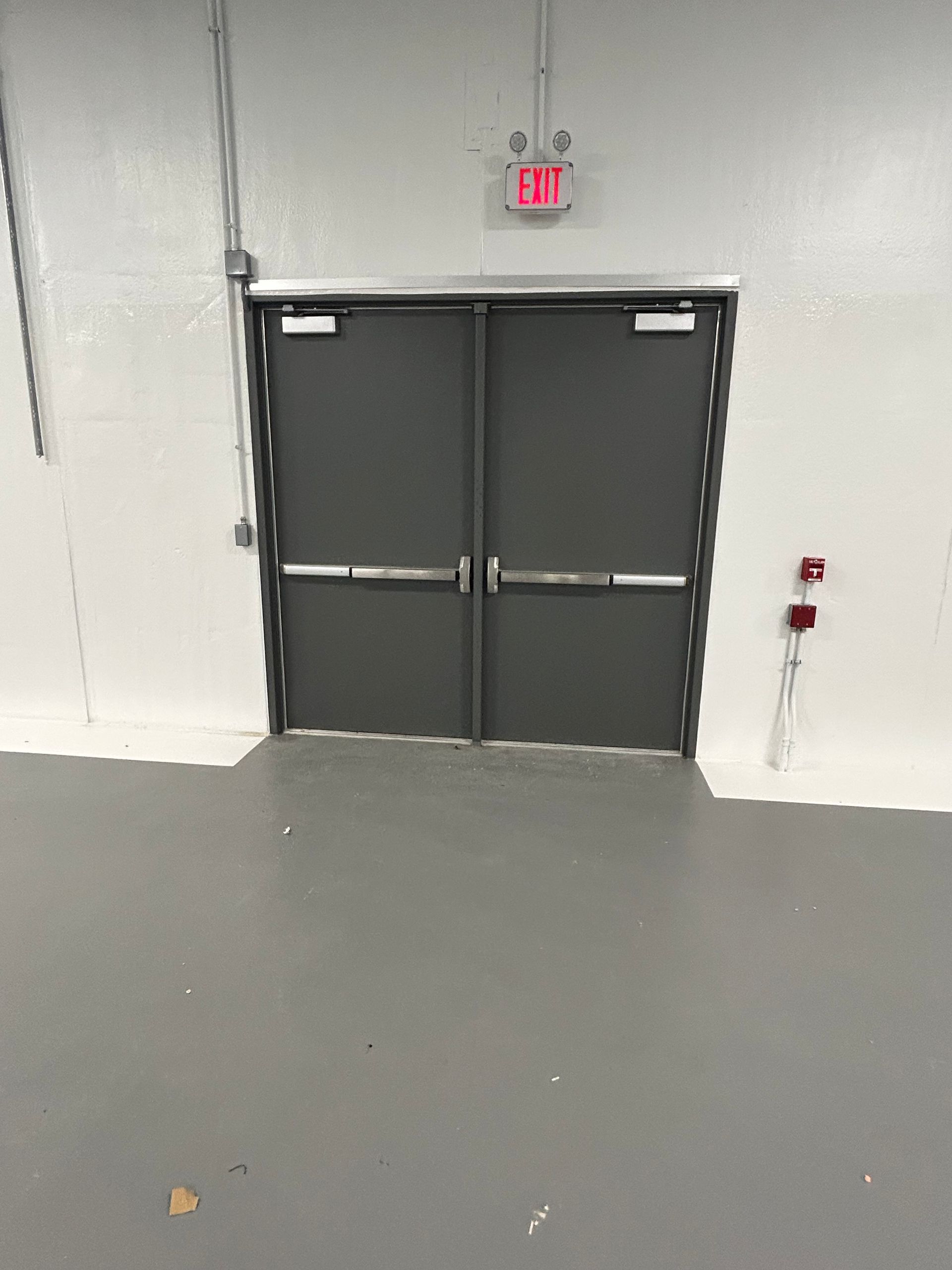 A gray double door with push bars and an EXIT sign above, set in a white and gray room.