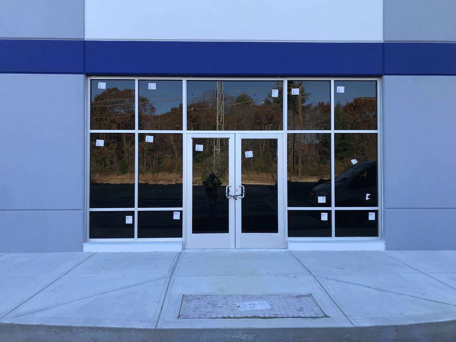 Exterior view of a building with glass doors and windows. Gray and blue facade, concrete sidewalk, and a door mat.