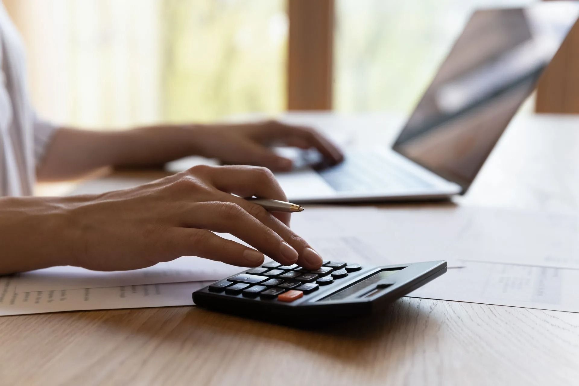 Person's hands using a calculator and laptop on a wooden desk, likely working on finances.