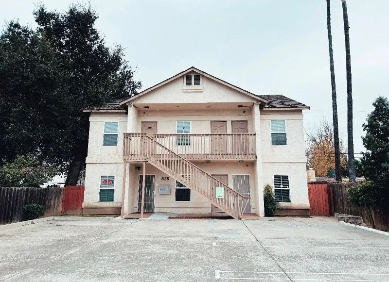 Two-story beige apartment building with wooden staircase and doors, on a concrete lot, overcast sky.