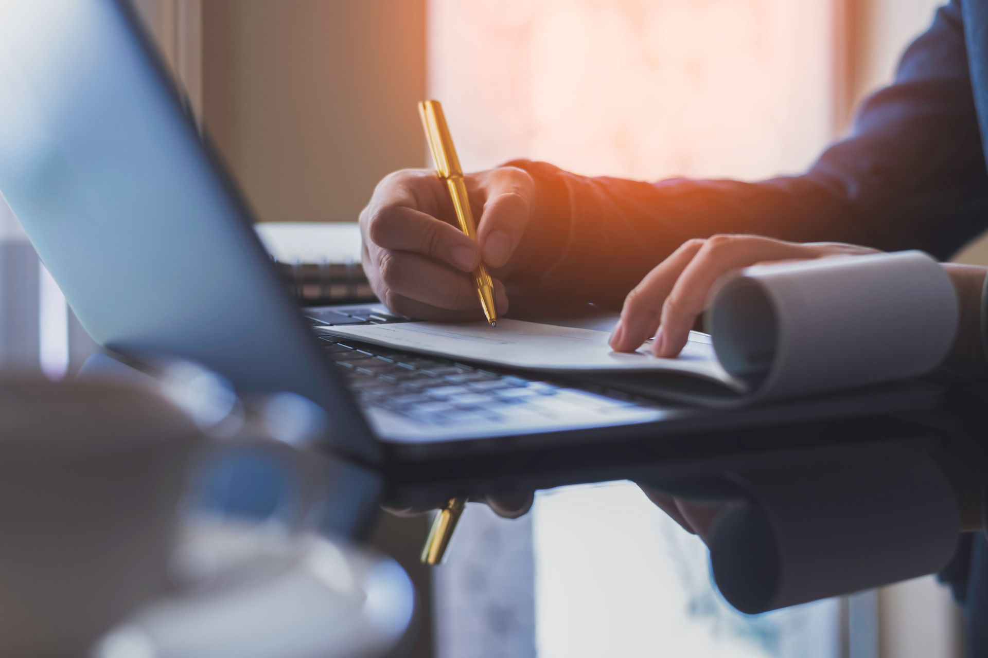 Person writing on paper with a pen next to a laptop; sunlight shining on desk.