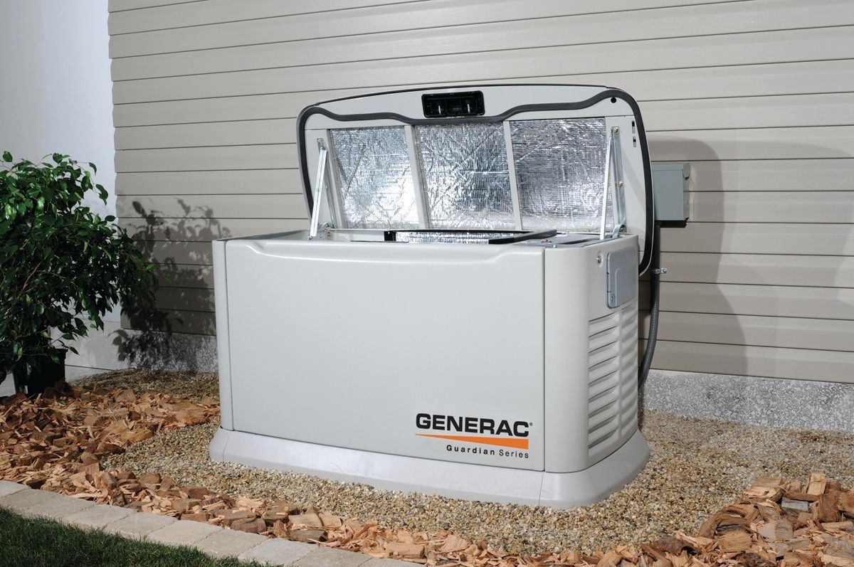 Generator with silver insulated cover beside a house on gravel, next to a bush and siding.