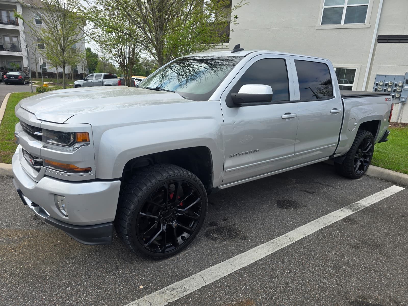 A silver Chevrolet Silverado pickup truck parked in an asphalt parking lot near an apartment building