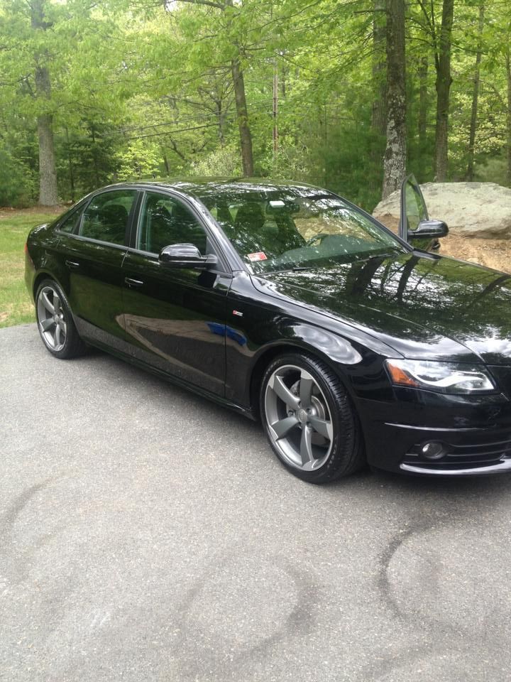 A black car is parked in a driveway with trees in the background.