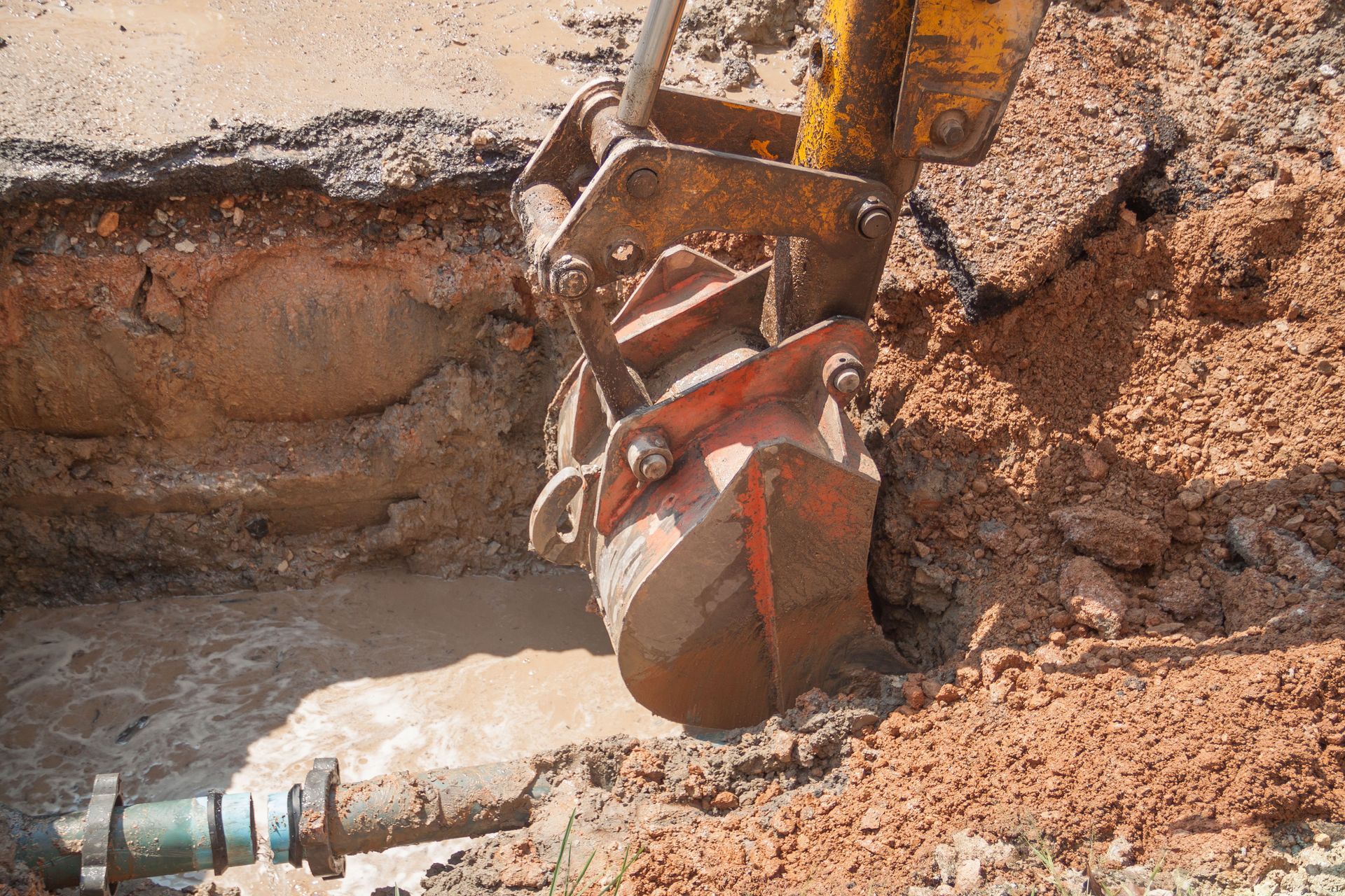 Excavator bucket digging in dirt near a broken water pipe, releasing water.