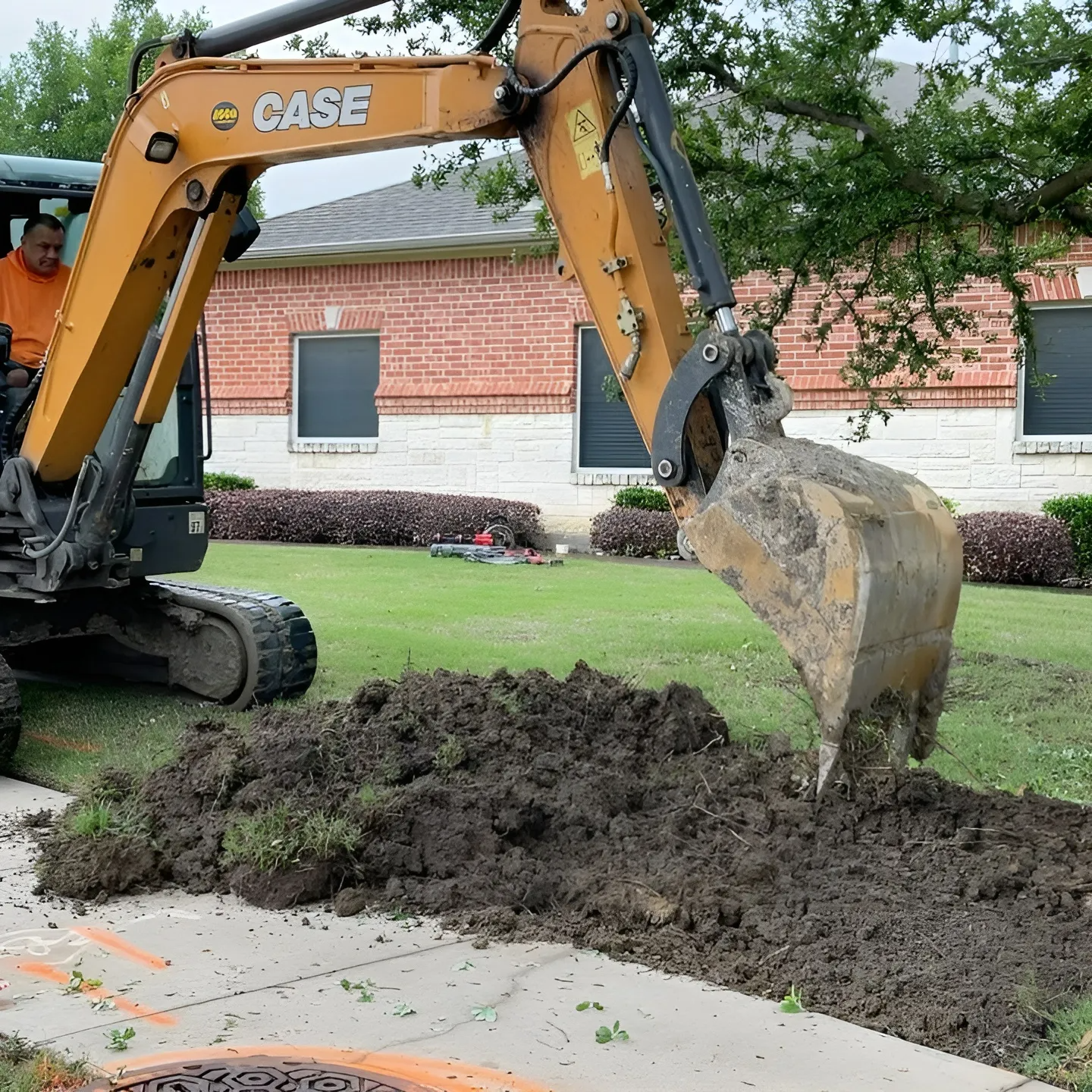 An excavator digging on a residential lawn, a pile of dirt beside it. A person is visible in the cab.