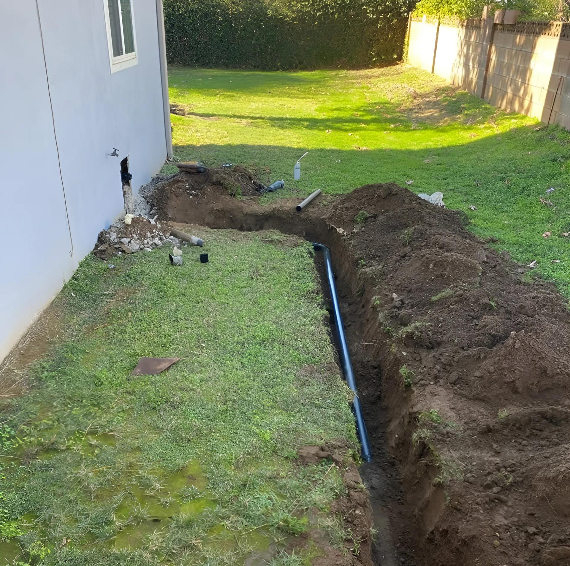 Trench dug beside a house, exposing black pipe. Green lawn, dirt pile, and cement wall visible.