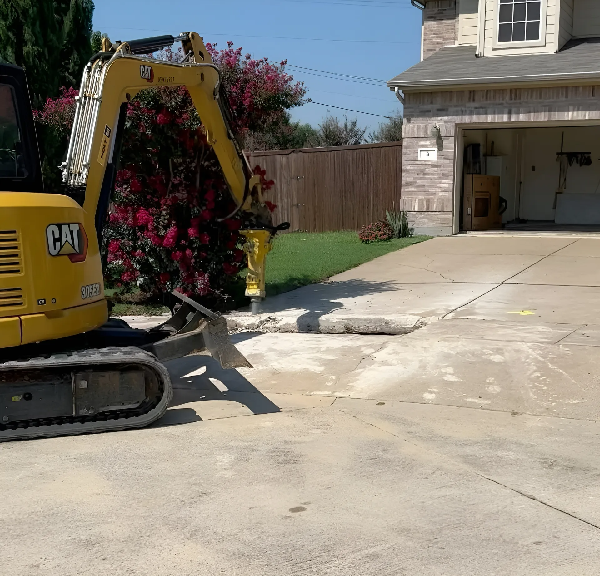 Yellow excavator breaking up concrete driveway in front of a house.