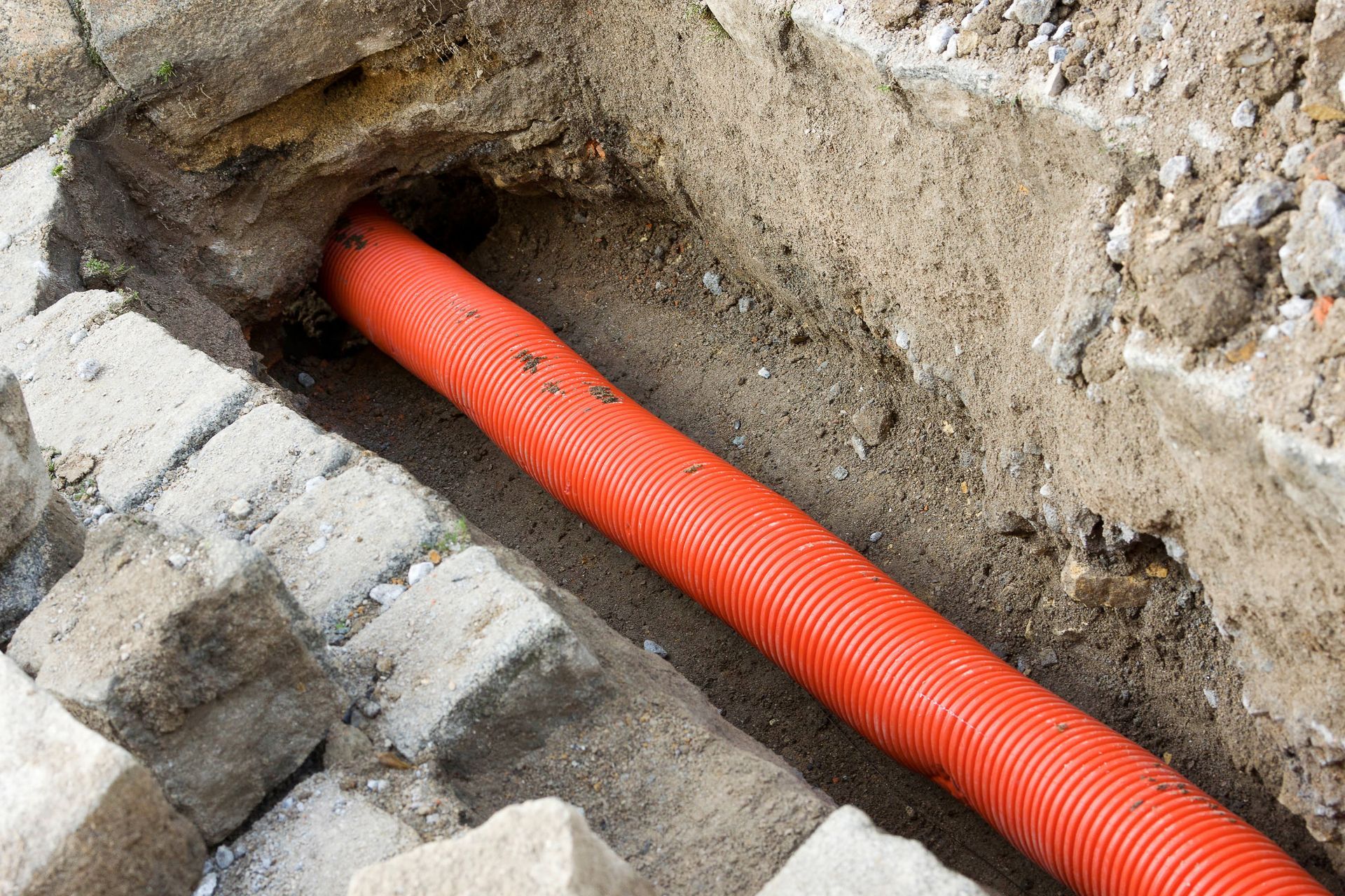 Orange corrugated conduit pipe in a trench, next to concrete blocks and earth.