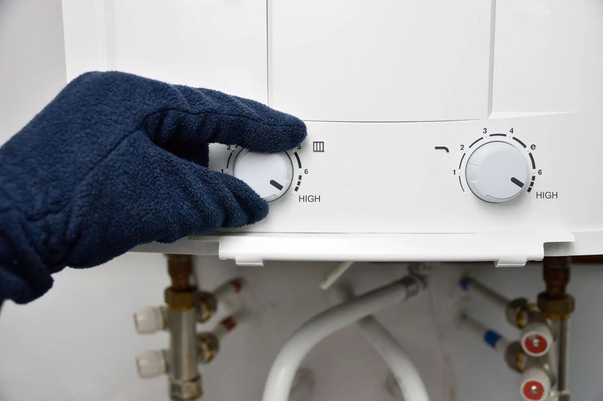 Gloved hand turning a dial on a white water heater, with pipes and valves visible below.