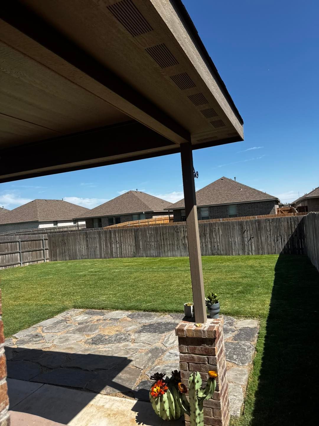 Backyard patio with a covered roof, brick pillar, and a view of houses and a fence on a sunny day.