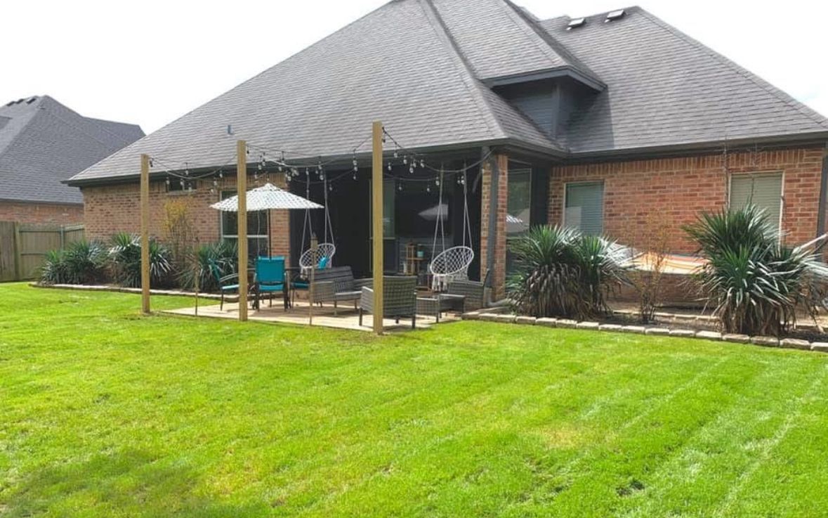 Backyard with brick house, patio with seating, and lush green lawn. Wooden posts support string lights.