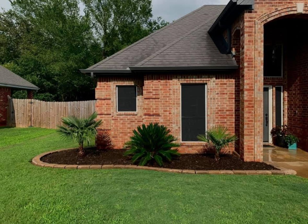 A brick house with a landscaped front yard, featuring a dark roof, two black window screens, and palm trees.