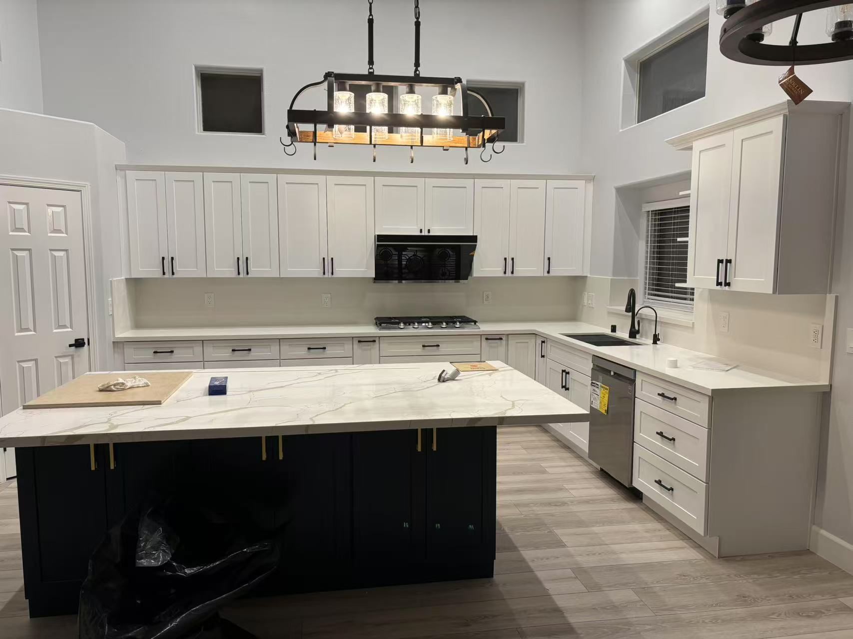 A modern kitchen with white upper cabinets, light countertops, a dark blue island, and a black chandelier.