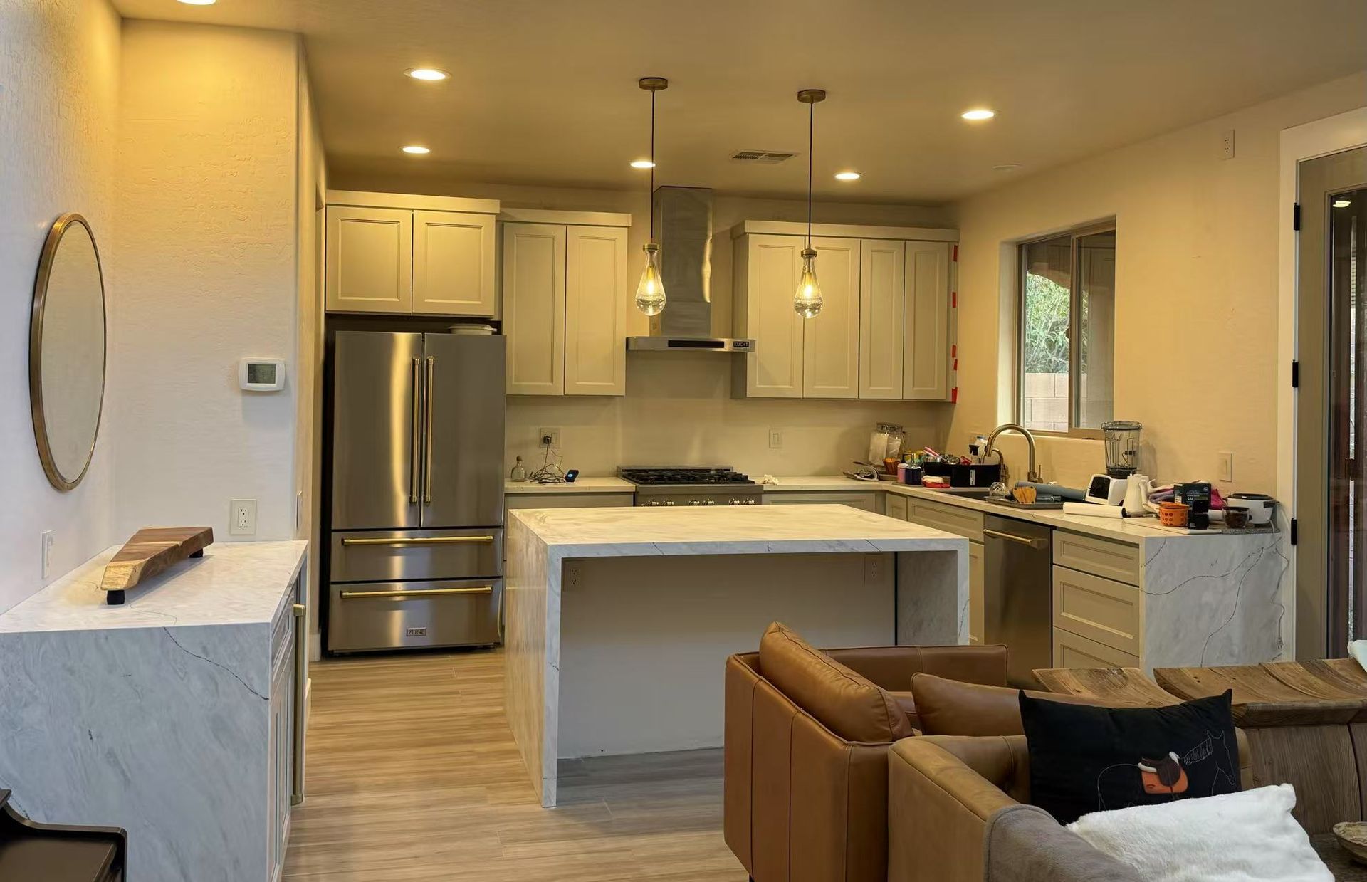 A modern kitchen with white cabinets, stainless steel appliances, a marble-topped island, and a beige sofa in the foreground.