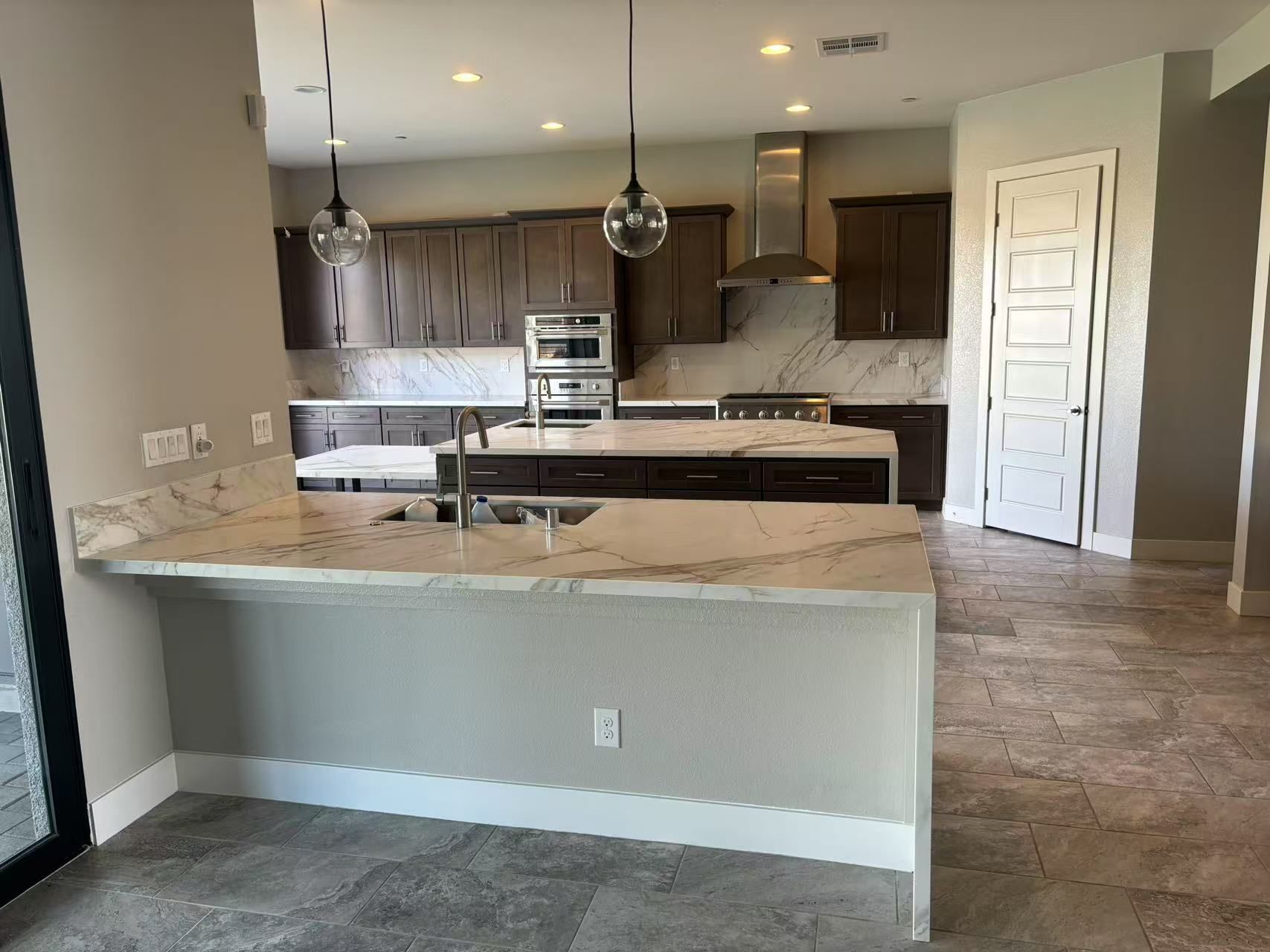 Modern kitchen with dark cabinets, marble-patterned countertops, two hanging pendant lights, and light tile flooring.