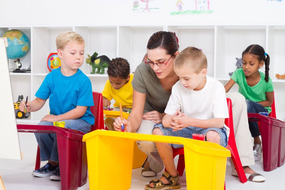 Teacher helping children paint at a table in a colorful preschool classroom.