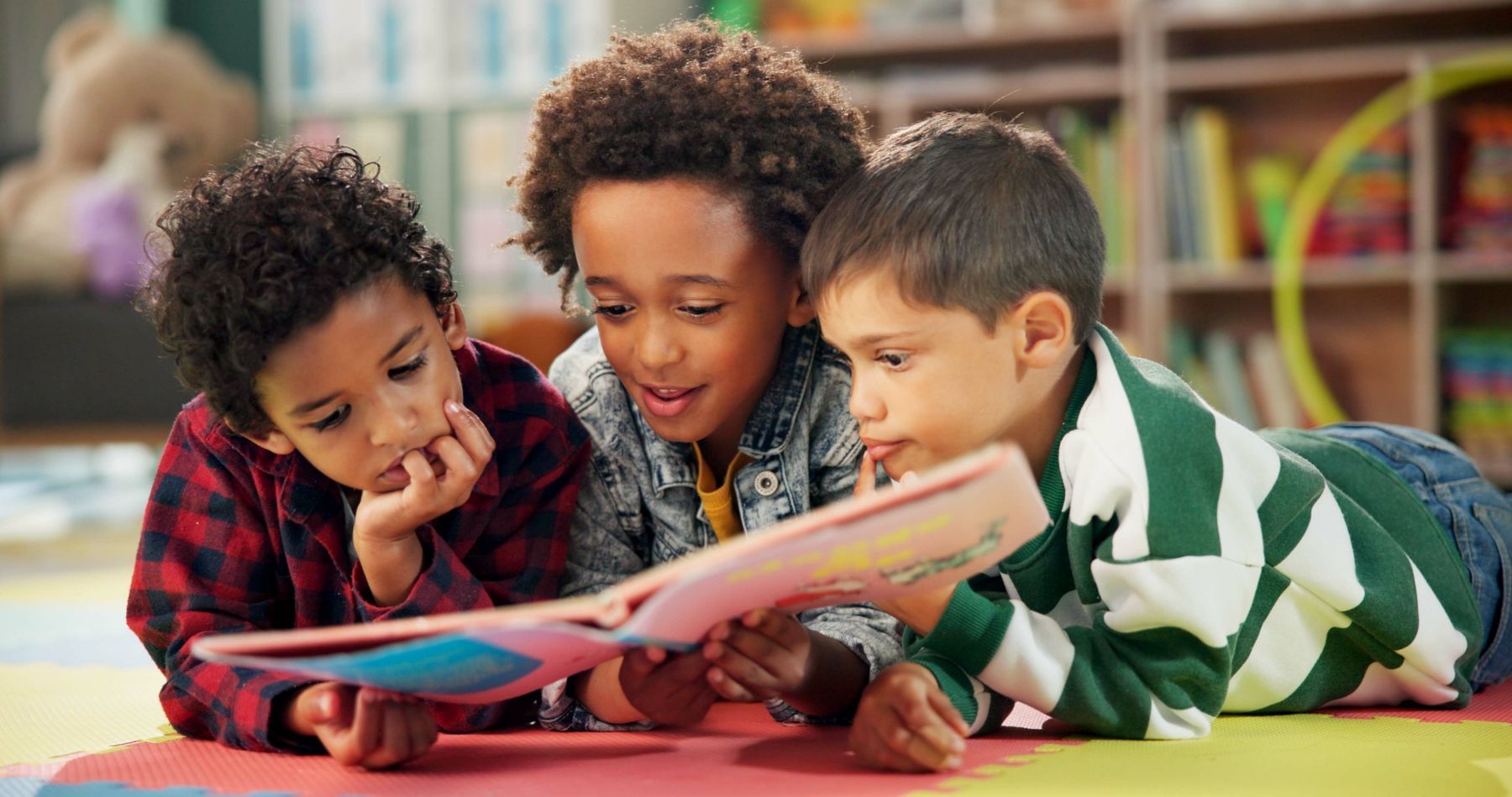 Three young boys are laying on the floor reading a book together.