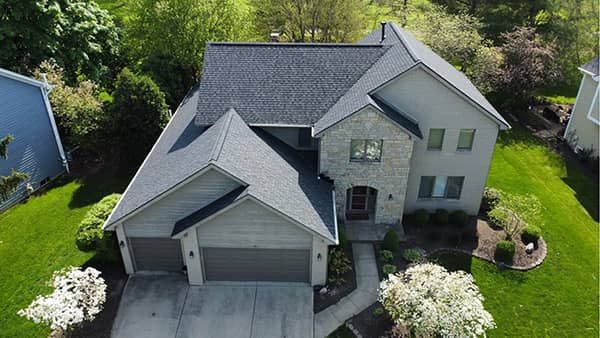 Two-story house with gray roof, stone facade, and green lawn, viewed from above on a sunny day.