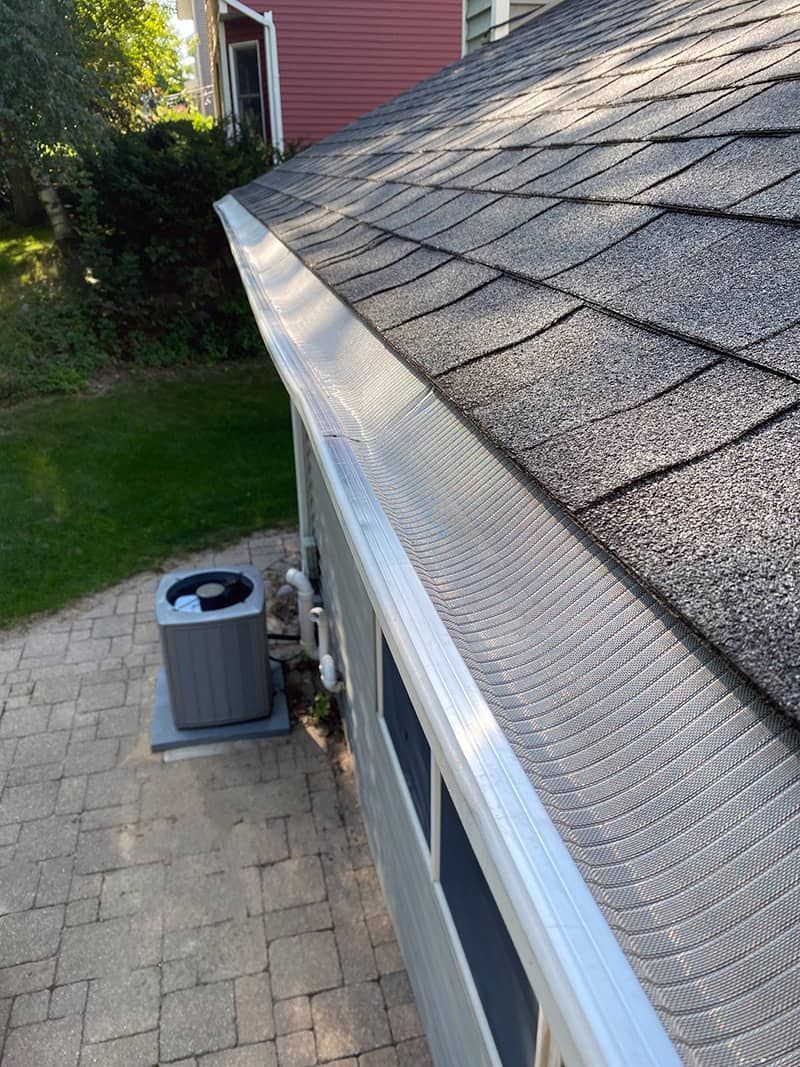 Silver gutter along a roof with gray shingles, an air conditioner, and a brick patio.