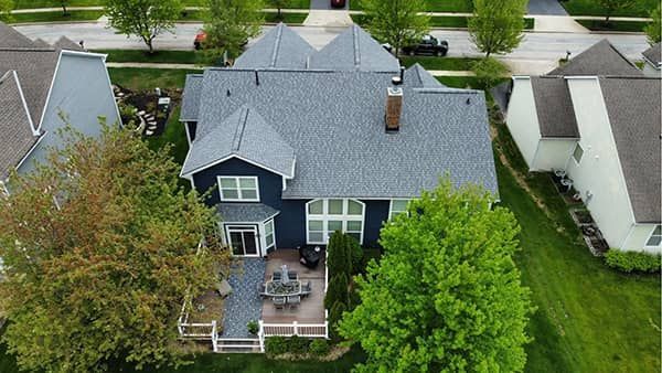 Overhead view of a blue-sided house with a gray roof and a wooden deck; surrounded by trees and green grass.