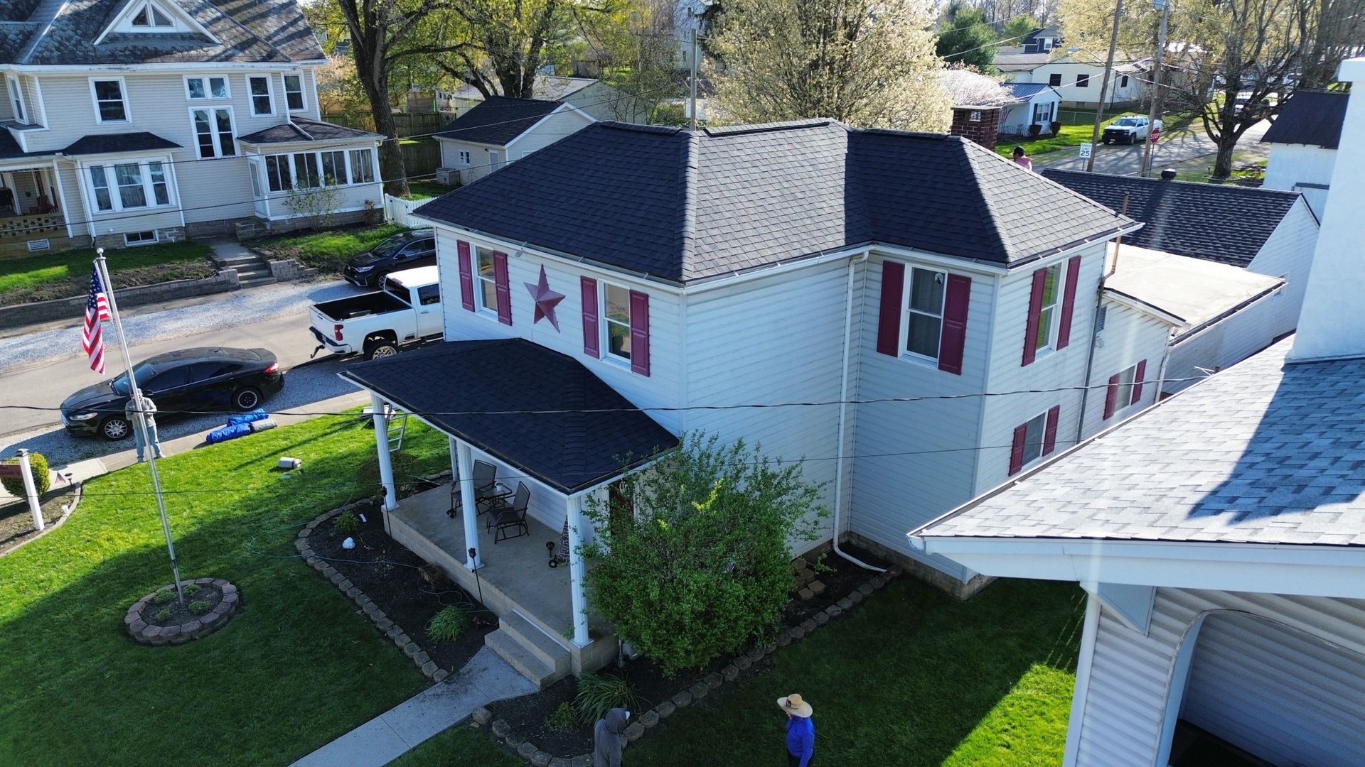 Two-story white house with black roof, red shutters, porch, and a lawn, in a town.