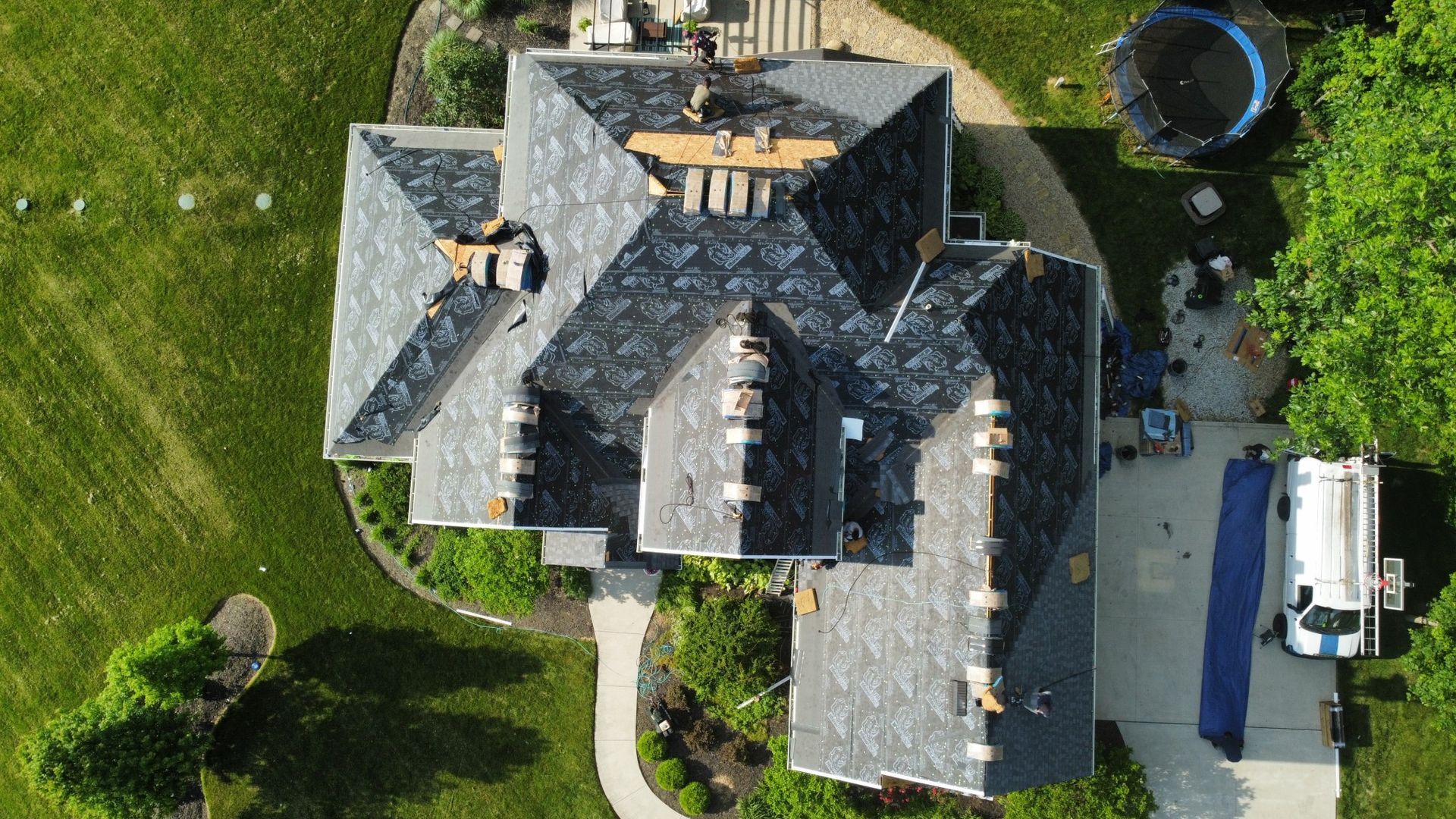 Aerial view of house with dark gray roof under renovation, surrounded by green lawn and trees.