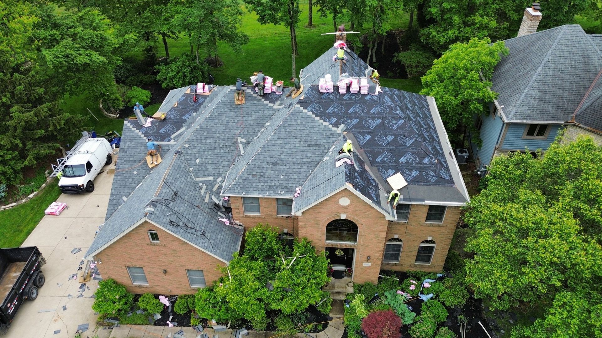 Roofers working on a two-story brick home; debris on ground, trees surround.
