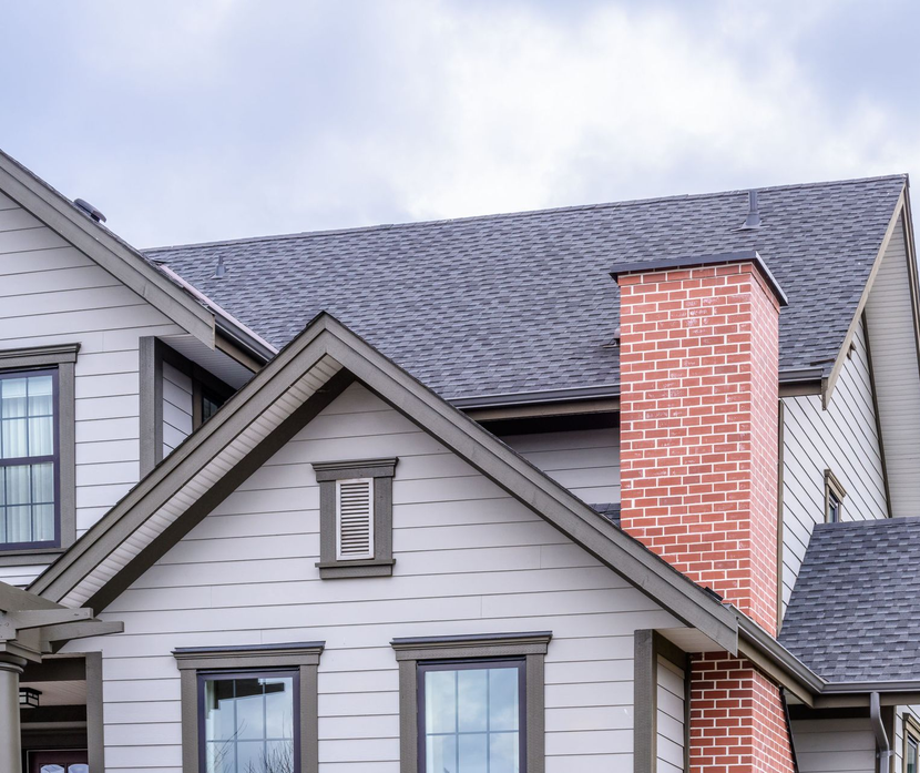 Gray house with dark roof and red brick chimney against a cloudy sky.