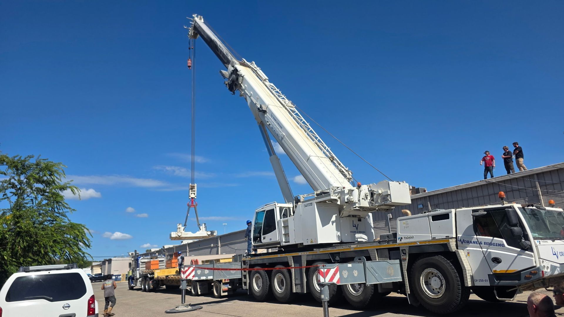 Crane lifting equipment beside a road under a clear blue sky, with workers on top and trucks below