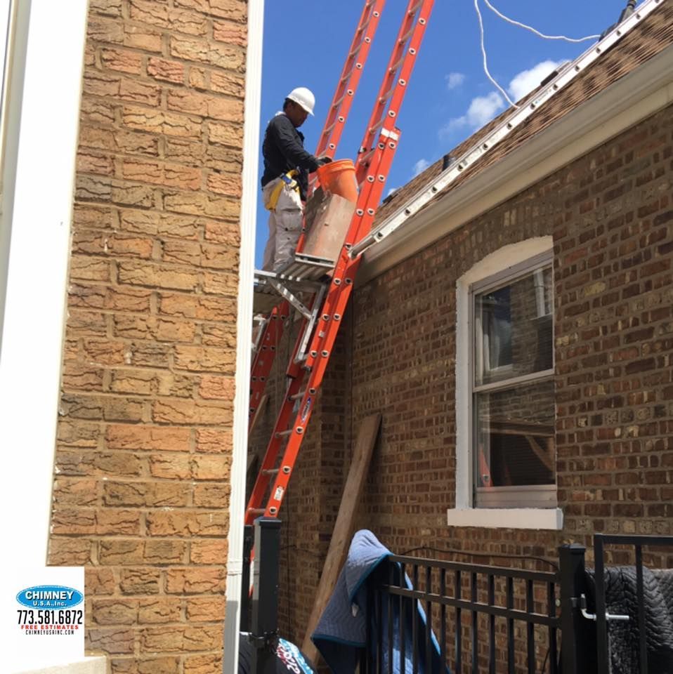 Man on ladder, brick building. He is painting near the roof, orange ladder, white hardhat, blue sky.