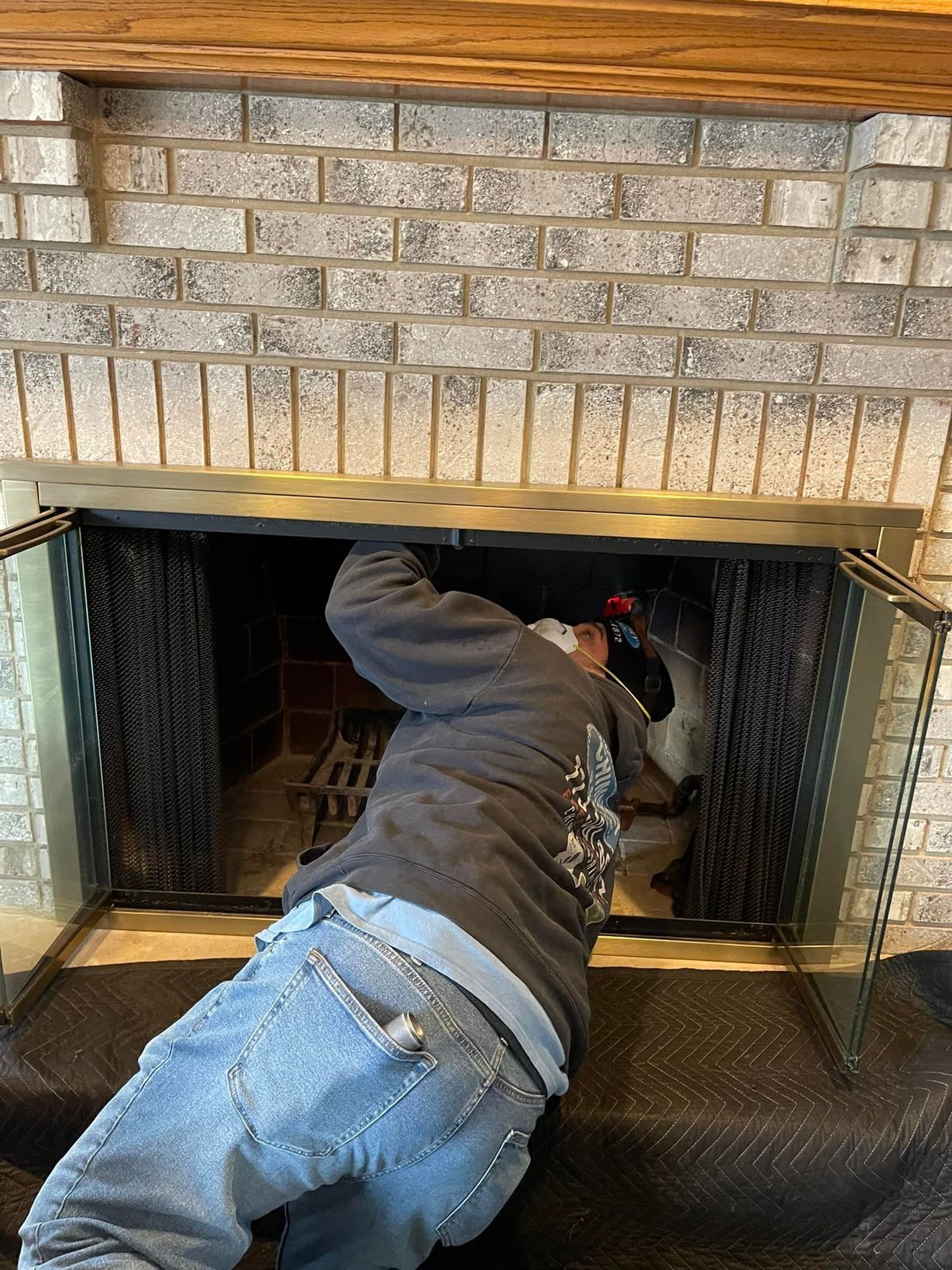 Man cleaning inside a brick fireplace with glass doors; blue jeans, gray jacket.
