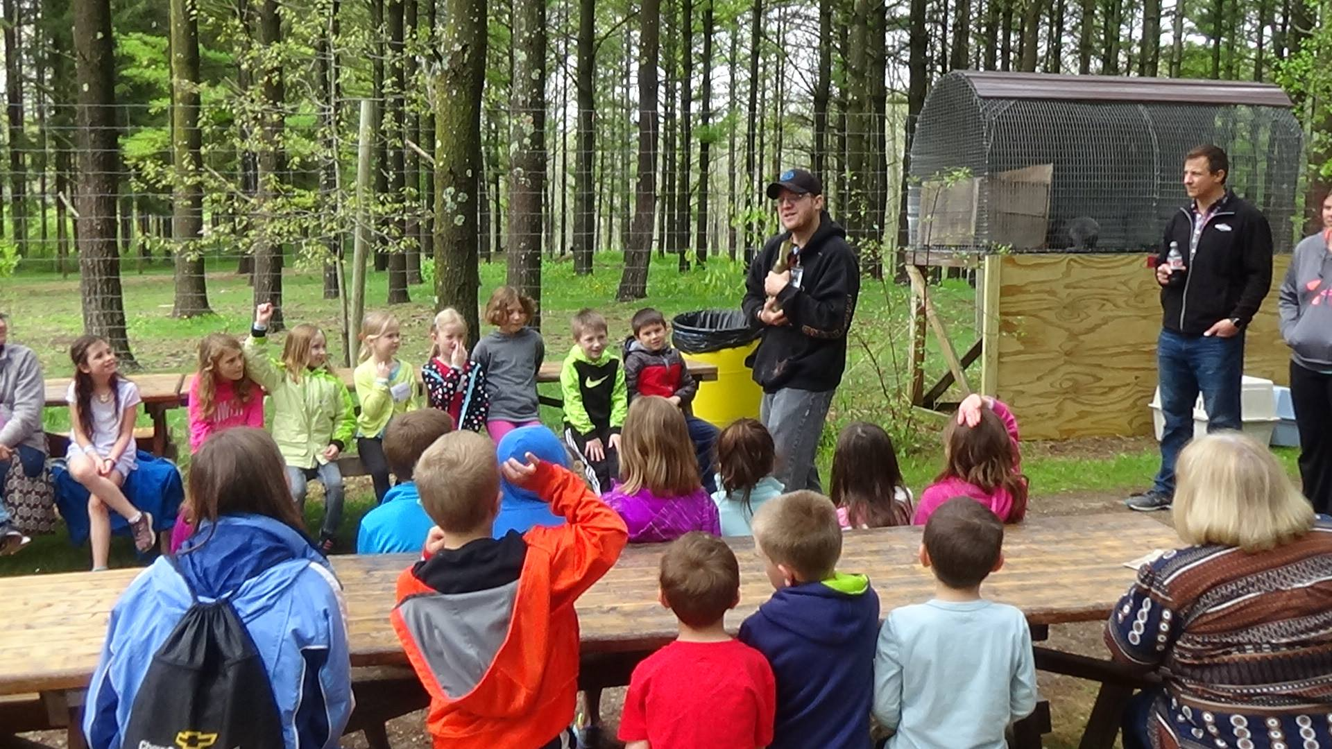 Children listening to a presentation outdoors. A man speaks, holding a trophy. Green trees surround a small structure.