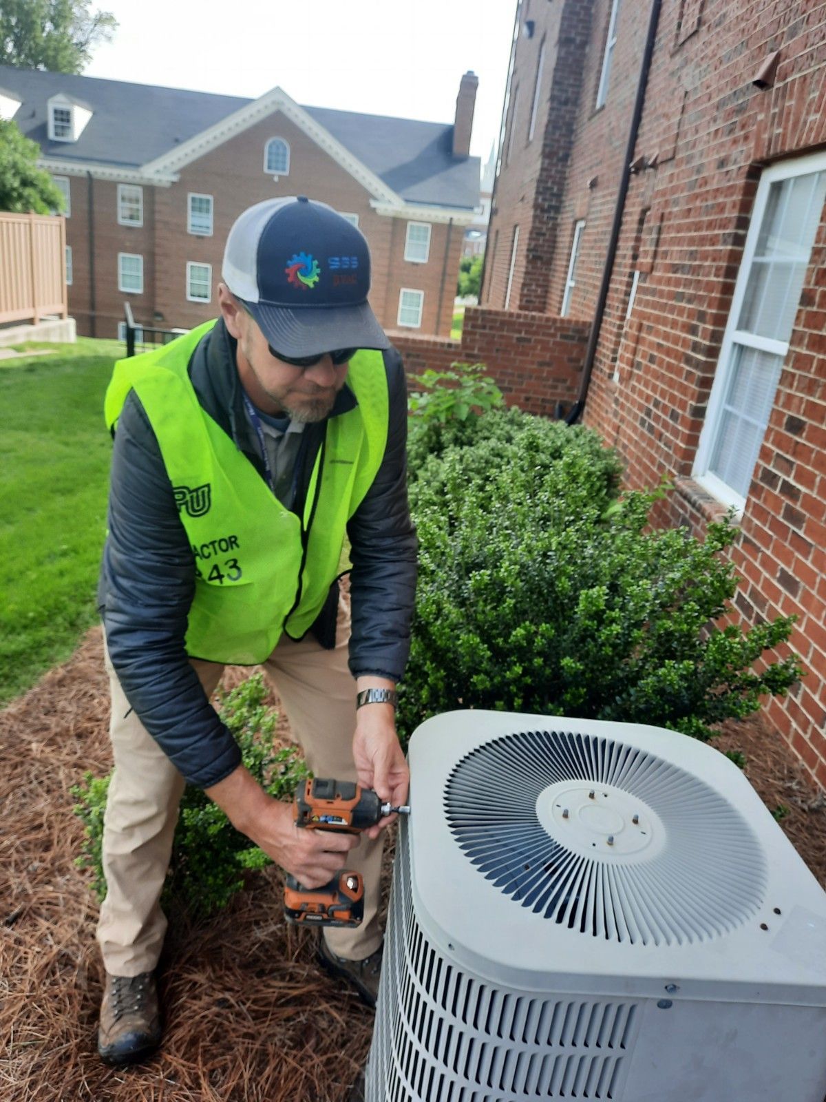 Technician in neon safety vest servicing an outdoor HVAC unit beside a brick building.
